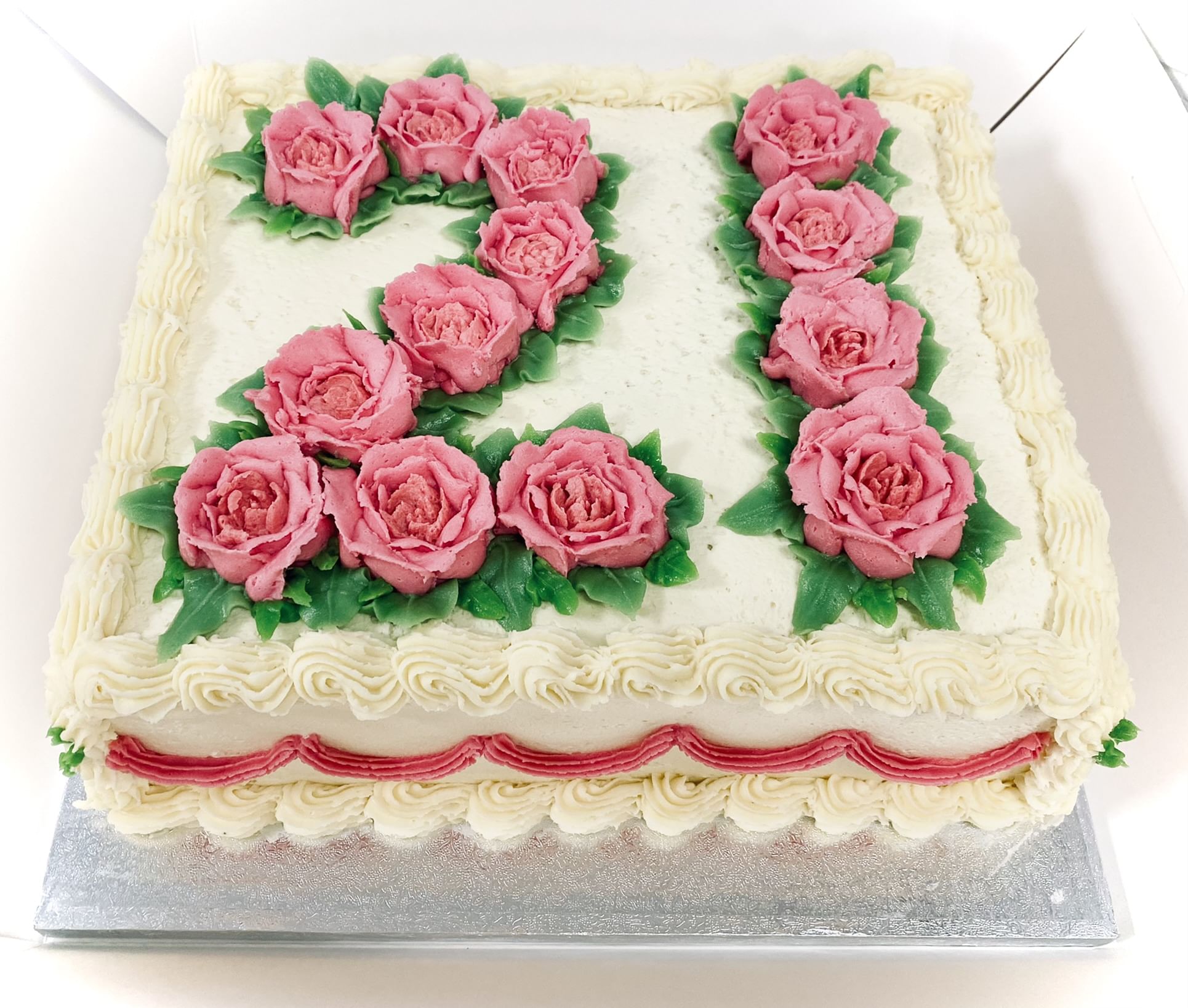 A square cake decorated with the number '21' made of pink flower-shaped icing. The cake is frosted with white icing and has piped white edges, with a border of white and pink icing at the base. The cake sits on a silver-colored board.